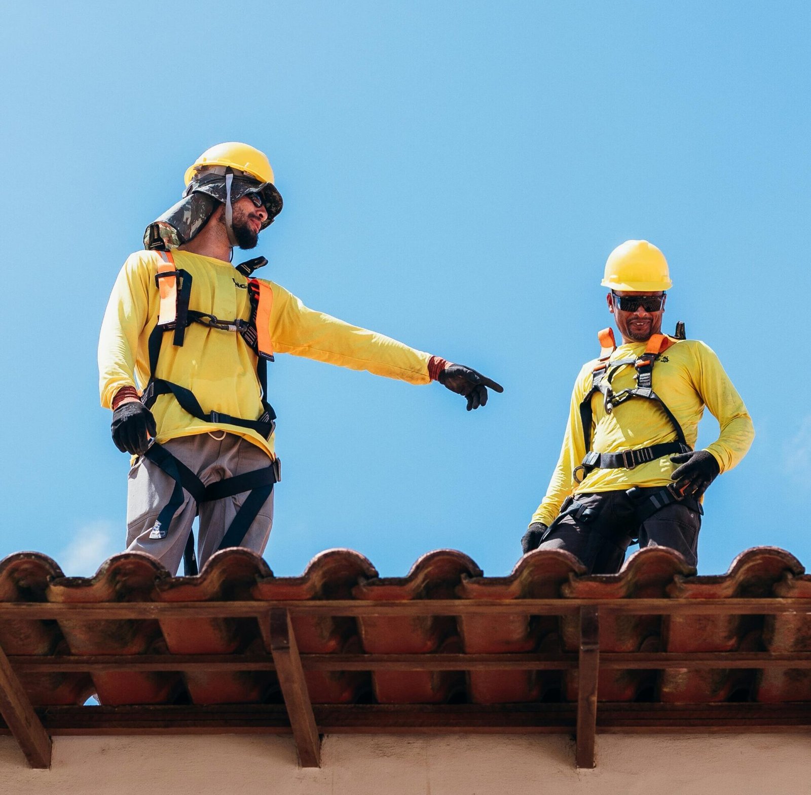 Two construction workers in safety gear discussing roofing project under clear blue sky.